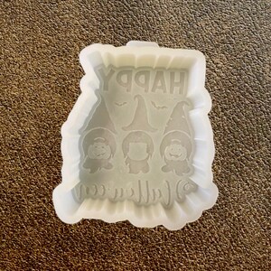 May include: A clear, flexible silicone mold for crafting. The mold features the words "HAPPY HALLOWEEN" and three gnome figures. The gnomes are wearing pointed hats. A bat graphic is also included in the design. The mold is designed for resin casting.