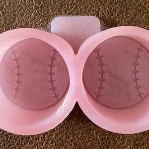 May include: Pink silicone mold with two baseball-shaped cavities. The mold is designed to create coasters or other small decorative items.