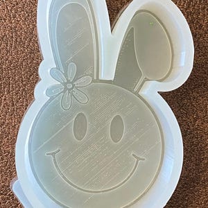 Puede incluir: Un molde de silicona transparente con forma de conejo con una flor y una cara sonriente. El molde está diseñado para hacer manualidades de resina.