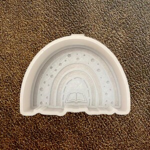 May include: A white, rainbow-shaped mold with an open book design in the center. The mold has a smooth surface and is designed for crafting. The rainbow has multiple layers with dotted details.