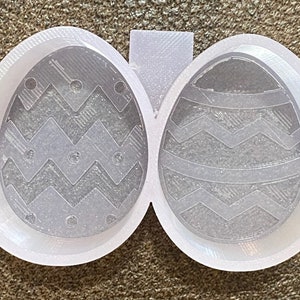 Pode incluir: Forma de plástico branco para fazer dois ovos de Páscoa. A forma tem um padrão de chevron em cada ovo.