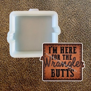 Puede incluir: Un molde cuadrado de silicona transparente con bordes redondeados y un pequeño asa. Junto a él, una pegatina marrón con el texto "I'M HERE FOR THE Wrangler BUTTS" en negro, con un borde cosido.