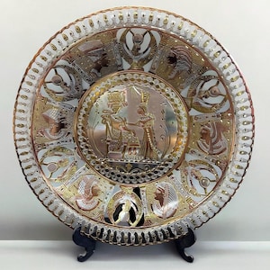 Puede incluir: Un plato redondo decorativo de metal con un diseño intrincado que presenta jeroglíficos egipcios y una escena de un faraón y una reina. El plato está hecho de cobre y plata y tiene un acabado dorado.