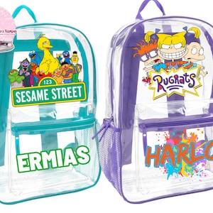 May include: Two clear backpacks with colorful cartoon characters. The blue backpack has Sesame Street characters and the text "Sesame Street" and "ERMIAS". The purple backpack has Rugrats characters and the text "Rugrats" and "HARLO".