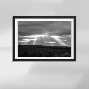 Può includere: Una fotografia di paesaggio incorniciata in bianco e nero che mostra un campo scuro ed esteso sotto un cielo drammatico e nuvoloso. Luminosi raggi solari squarciano la fitta coltre di nubi, proiettando luce verso l'orizzonte in una scena.
