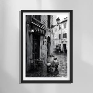 Può includere: Una stampa artistica incorniciata in bianco e nero che raffigura una scena di strada europea con uno scooter parcheggiato su un sentiero di ciottoli bagnato. È visibile un caffè con l'insegna CAFFE, sedute all'aperto e lampade da parete.