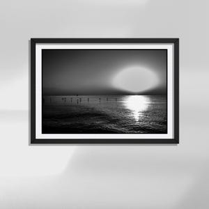 Può includere: Una fotografia di paesaggio in bianco e nero incorniciata che ritrae un tramonto su uno specchio d'acqua calmo. Il sole è posizionato basso all'orizzonte, proiettando un riflesso luminoso sulla superficie increspata, con piccole sagome visibili.
