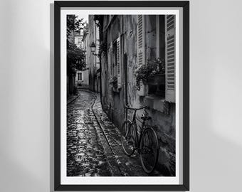 Puede incluir: Una impresión de arte mural enmarcada en blanco y negro que representa un callejón estrecho y húmedo de adoquines en una ciudad europea. Una bicicleta está aparcada contra una pared de piedra desgastada con ventanas con contraventanas.