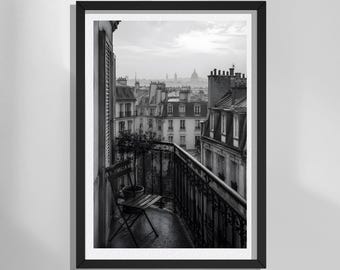 May include: A black and white framed wall art print depicting a balcony view overlooking a city skyline with historic buildings. A small metal chair sits on the balcony, which features a decorative iron railing and a potted plant in the corner.