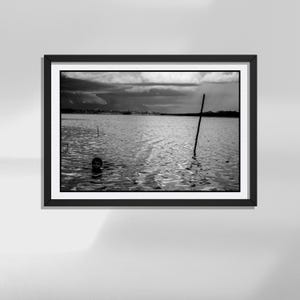 Può includere: Una fotografia incorniciata in bianco e nero che ritrae la testa di una persona parzialmente immersa in un grande specchio d'acqua. Un palo di legno alto e sottile si erge verticalmente nell'acqua sulla destra, con una costa lontana sotto un cielo nuvoloso.