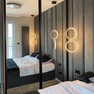 Könnte beinhalten: Ein modernes Schlafzimmer mit einem grauen, gepolsterten Kopfteil und zwei Spiegeltüren. Die Schranktüren spiegeln das Schlafzimmer wider, einschließlich eines Bettes mit weißen Bettwäsche und schwarzen Kissen. Drei runde Pendelleuchten hängen von der Decke und erhellen den Raum.