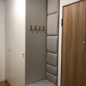 Puede incluir: Un armario blanco con un banco acolchado gris y una puerta de madera. El banco tiene un compartimento de almacenamiento debajo. Hay ganchos de madera en la pared sobre el banco.