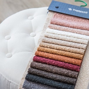 Może przedstawiać: Książka próbek tkanin z różnymi kolorami i fakturami. Książka jest oznaczona "Toptextil" i jest prezentowana na białym pufie.