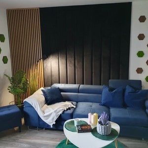 Op de afbeelding: Een blauwe hoekbank met een wit plaid en kussens in een woonkamer met een houten lattenwand, een zwarte muur met verticale panelen en een witte muur met groene mos hexagon wanddecoratie.