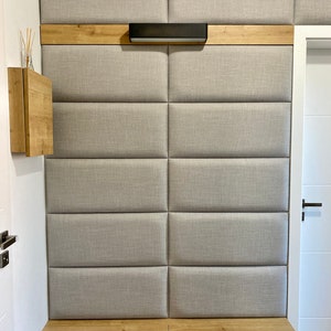 Puede incluir: Un panel de pared tapizado gris con un estante de madera y una lámpara negra. El panel está dividido en secciones rectangulares y tiene un diseño acolchado. El estante está hecho de madera clara y tiene un diseño simple. La lámpara tiene una forma rectangular larga y está montada en el estante. El panel de pared está montado en una pared blanca y es parte de una habitación más grande.