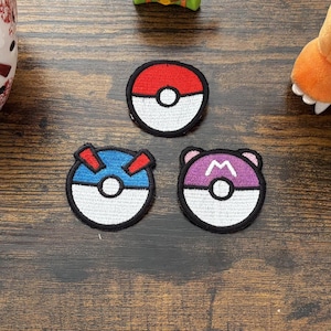 May include: Three circular embroidered patches featuring designs inspired by Poke Balls. One patch is red and white, one is blue and white with red accents, and one is purple and white with a black M symbol. The patches are arranged in a triangular formation.