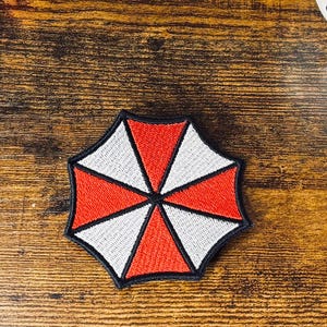 May include: An embroidered patch shaped like an umbrella with alternating red and white triangular segments outlined in black. Nearby are two spools of thread, one black and one red, and a white ceramic mask with black and red painted details.