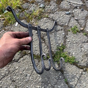 May include: Hand-forged, dark metal wall hook with three curved hooks. The hook has a decorative, curved top and is held by a hand against a textured gray surface. The hooks are connected by hinges.