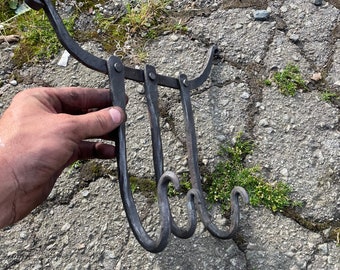 May include: Hand-forged, dark metal wall hook with three curved hooks. The hook has a decorative, curved top and is held by a hand against a textured gray surface. The hooks are connected by hinges.