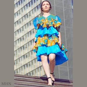 Puede incluir: Una mujer con un vestido estampado en azul y amarillo con una falda con volantes. El vestido tiene un escote alto y está decorado con adornos dorados. La mujer está de pie en una escalera.