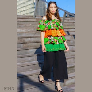 May include: A woman wearing a green, orange and white off-the-shoulder top with a ruffled hem and black trousers. The top has a floral print and is layered with different fabrics.