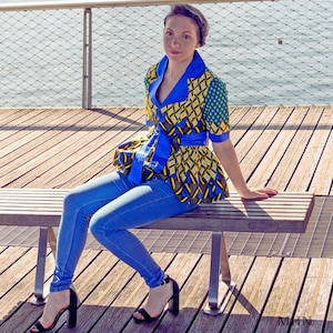 Puede incluir: Una mujer lleva una blusa con estampado azul y amarillo con una faja azul atada a la cintura. Está sentada en un banco de madera con las piernas cruzadas y lleva pantalones vaqueros azules y tacones negros.