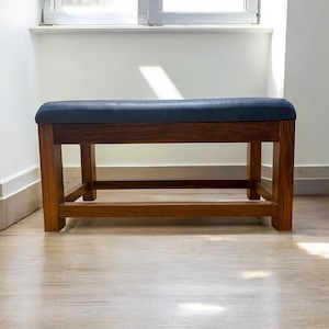 May include: A wooden bench with a dark blue cushioned seat. The bench has a rectangular shape with a wooden frame and legs. The bench is placed in front of a window, with natural light illuminating the scene.
