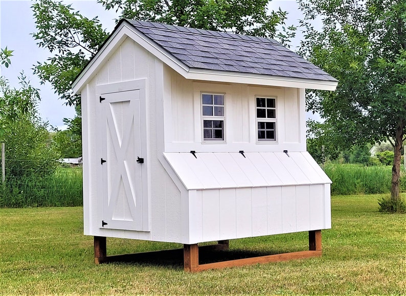 May include: White wooden chicken coop with a black door and two windows. The coop has a black shingled roof and is raised on four wooden legs.