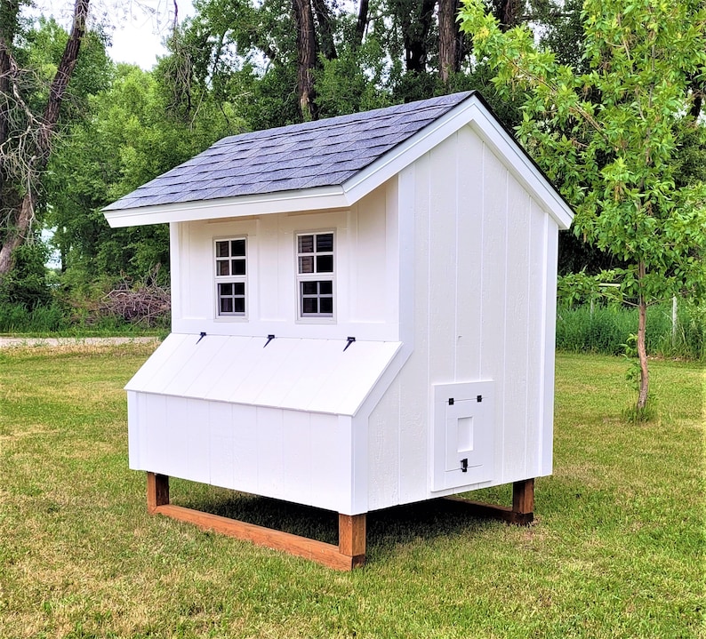 May include: White wooden chicken coop with a black shingled roof. The coop has two windows, a nesting box, and a door. The coop is sitting on a wooden base.