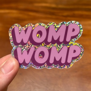 May include: A pink and purple glitter sticker with the text "WOMP WOMP" in a bubble font.