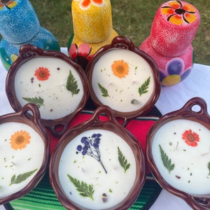 May include: Five brown ceramic candle holders with white candles and dried flowers. The candles are decorated with dried flowers and greenery. The candle holders are sitting on a colorful Mexican blanket.