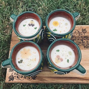 May include: Four green and brown ceramic mugs with handles, each filled with a white candle and decorated with dried flowers. The mugs are arranged on a wooden surface.