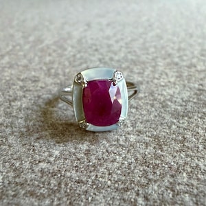 Könnte beinhalten: Ein silberfarbener Ring mit einem großen, kissenförmigen, tief pink-roten Edelstein in der Mitte. Der Stein ist von einer glatten, hellen, schimmernden Umrandung eingefasst und mit vier kleinen, klaren, rund geschliffenen Steinen an den Ecken verziert.