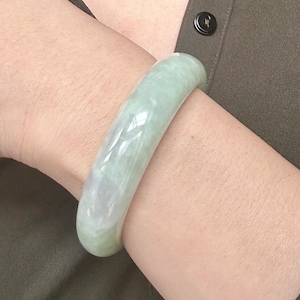 Op de afbeelding: Een gladde, gepolijste, cilindrische armband gemaakt van lichtgroene jade. De steen heeft een doorschijnende kwaliteit met subtiele witte, wolkachtige insluitingen en een hoogglanzende afwerking die het licht over het oppervlak weerkaatst.