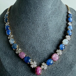 May include: A necklace featuring a series of oval cabochon-cut blue, pink, and white gemstones set in gold-toned metal. The stones are interspersed with small, flower-shaped clusters of clear, sparkling stones, creating a continuous link design.
