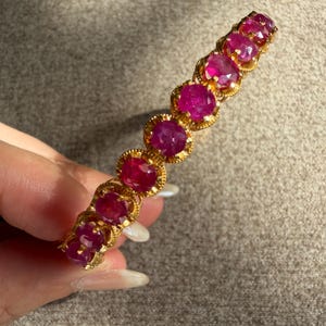 Puede incluir: Una pulsera de oro con una serie de piedras preciosas de corte ovalado en color rojo rosado intenso, engastadas en biseles de oro festoneados individuales. Las piedras están dispuestas en una sola fila, creando una línea continua de color.