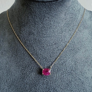 May include: A delicate gold chain necklace featuring a single, faceted, cushion-cut deep pink gemstone pendant. The stone is secured in a simple four-prong setting, suspended centrally from a thin, polished gold-tone metal link chain.
