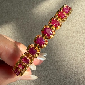 Puede incluir: Una pulsera de oro con una serie de rubíes de color rojo rosado intenso, de talla ovalada, engastados en monturas individuales de oro festoneado. Las piedras están dispuestas en una sola fila, aseguradas por garras en el marco de oro amarillo.