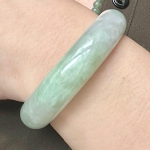 May include: A smooth, cylindrical bangle bracelet crafted from pale green jadeite with subtle white cloud-like inclusions. The polished surface of the bangle reflects light, showcasing a translucent appearance and a rounded, continuous circular form.