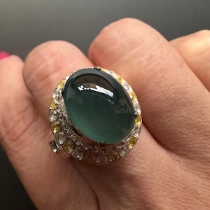 May include: A large, oval-shaped ring featuring a dark teal gemstone. The gemstone is surrounded by a halo of small, white and yellow stones set in a silver-colored metal band. The ring is shown on a person's finger.