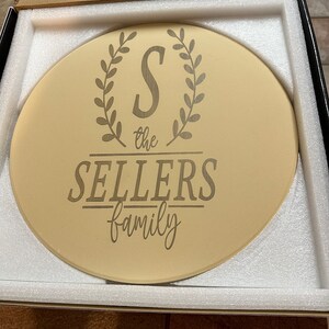Op de afbeelding: Een ronde, beige decoratieve plaat met de gegraveerde tekst "S the SELLERS family". De letter "S" staat in een lauwerkrans. De plaat wordt gepresenteerd in een zwarte doos met witte schuimverpakking.