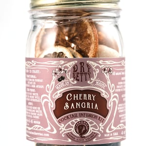May include: A glass jar with a gold lid filled with dried fruit and spices. The label reads "Dry Betty Cherry Sangria Cocktail Infusion Kit" and "Long Live Lilibet".
