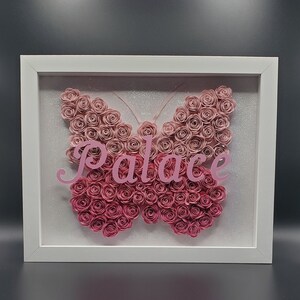 Könnte beinhalten: Ein weißer Rahmen mit einem rosa Papier-Schmetterling aus vielen kleinen Papierrosen. Der Schmetterling trägt den Namen "Palace" in rosa Buchstaben.