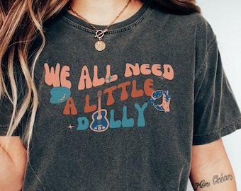 May include: Dark gray t-shirt with the text "WE ALL NEED A LITTLE DOLLY" in retro-style lettering. The design includes a cowboy hat, guitar, and other graphics in shades of orange, blue, and pink. The shirt is a casual, comfortable style.