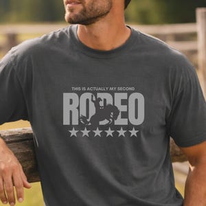 May include: Dark gray t-shirt with the text "THIS IS ACTUALLY MY SECOND RODEO" in gray. The word "RODEO" features a silhouette of a rider on a horse, with five stars below. The person is wearing blue jeans and a brown belt.