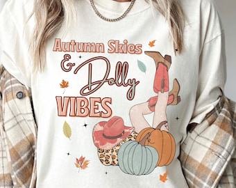 May include: White t-shirt with the text "Autumn Skies & Dolly Vibes" and a cartoon illustration of a woman wearing cowboy boots and a hat, sitting on a pumpkin and a leopard print pillow.