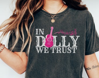 May include: Dark gray t-shirt with the text "IN DOLLY WE TRUST" in white, featuring a pink guitar graphic. Musical notes in pink are above the guitar. The shirt is a vintage style.