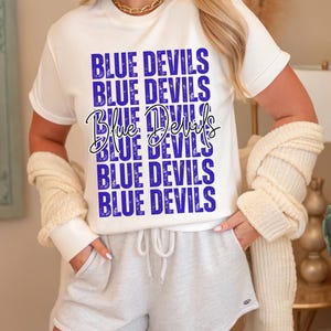 May include: A white t-shirt with the words "BLUE DEVILS" in a blue, stacked design. The words are repeated multiple times. The shirt is paired with gray shorts and a cream-colored cardigan.