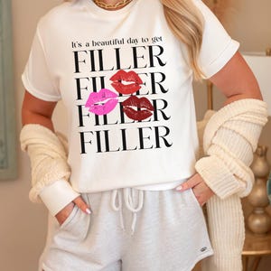 May include: White t-shirt with the text "It's a beautiful day to get FILLER" in black, with three sets of lips in red, pink, and burgundy. The person is wearing gray shorts and a gold necklace.
