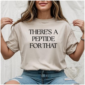 May include: A beige short-sleeved t-shirt featuring the black text THERE'S A PEPTIDE FOR THAT printed in a bold, serif font across the center of the chest. The garment has a crew neckline and a relaxed fit, constructed from a solid-colored fabric.